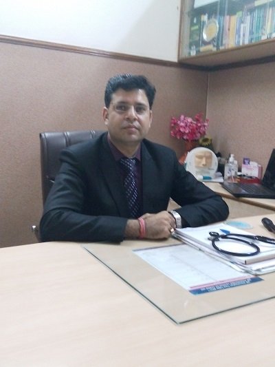 Dr. Vivek Goswami sitting at his desk in his office, wearing a black suit and tie, with a stethoscope and medical documents on the desk.